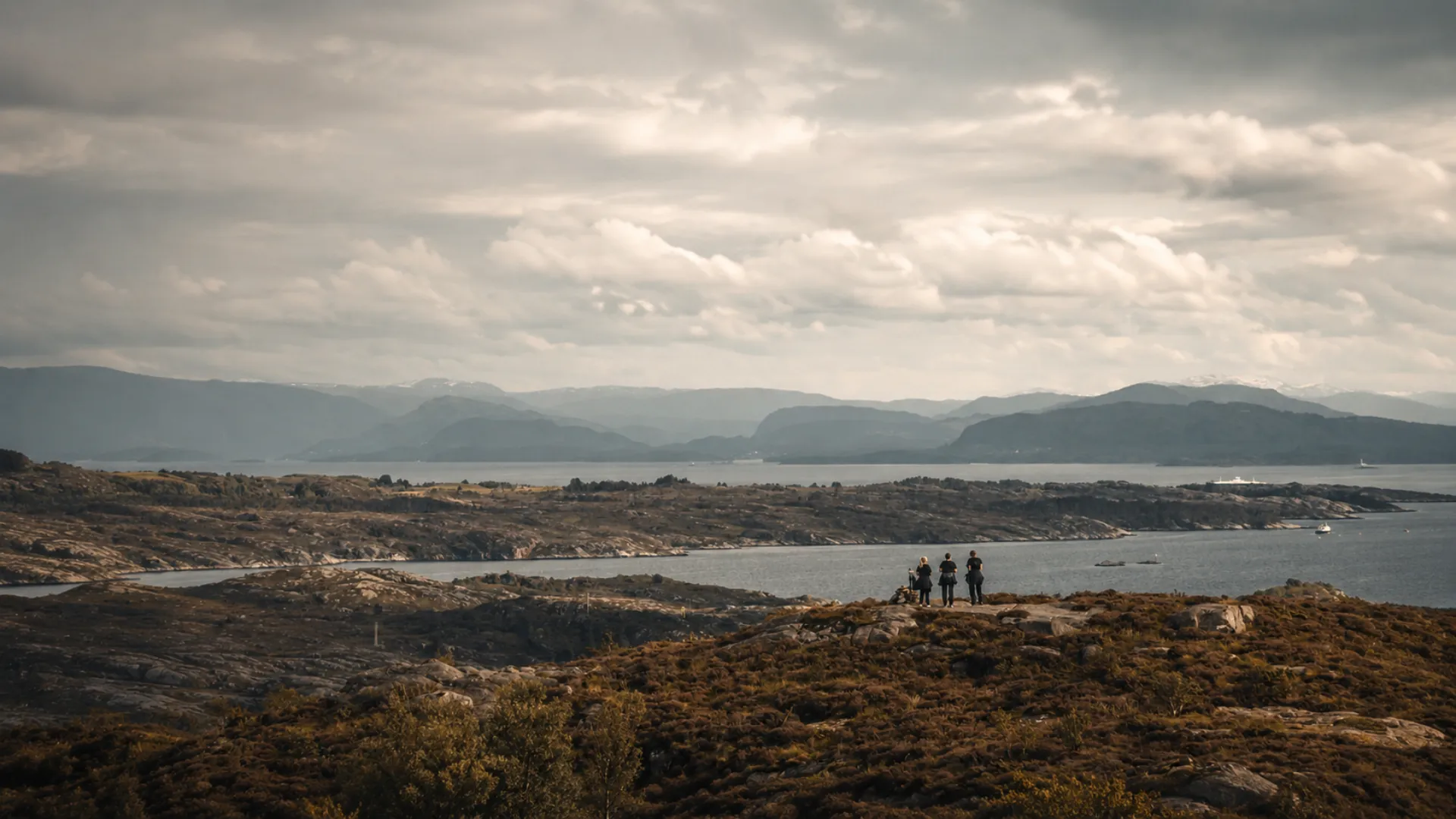Tre personer på en lyngkledd knaus med utsikt over øyer, fjord og fjell